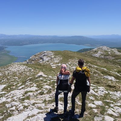 Blick von einem felsigen Karst-Gipfel der Dinara hinunter auf das weite, tiefblaue Buško jezero im Tal. Im Vordergrund stehen Martina und Martin von hinten am Gipfel, beide mit Wanderrucksack. Martina trägt ein rosa Kopftuch und einen weiss-schwarzen Rucksack, Martin einen grauen Hut und einen gelb-grünen Rucksack. Vor ihnen fällt der Hang in einen grasigen Karst-Sattel ab, dahinter erstreckt sich der See mit angedeuteten Halbinseln, im Hintergrund eine Kette flacher, dunstig-blauer Berge. Über allem ein wolkenloser, strahlend blauer Himmel.