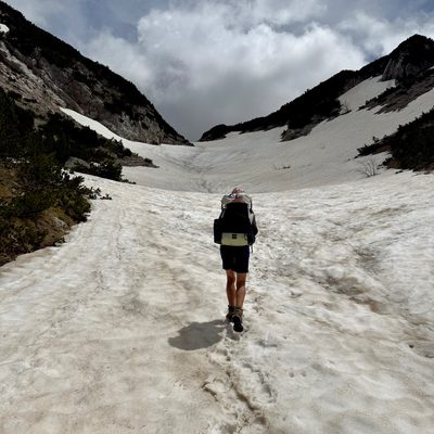 Eine Person mit grossem Wanderrucksack geht von hinten gesehen einen schmalen Schneepfad durch ein steiles, weisses Schneefeld in einem felsigen Bergkessel hinauf. Der Wanderer trägt kurze Hose, T-Shirt und Trekkingstiefel. Links und rechts steigen graue, felsige Karststeilhänge auf, teils noch mit Schneefeldern, teils mit grünen Pflanzen-Polstern bewachsen. Im Hintergrund ein zerklüfteter Bergrücken vor dunkel verhangenem Wolkenhimmel. Im Schnee sind die Spuren früherer Wanderer als Fussabdrücke zu sehen.