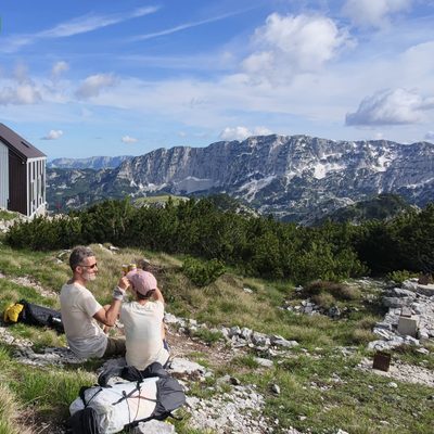 Blick von einem grasigen Karstplateau auf der Dinara auf den Planinarski Dom Vilinac, eine kleine dunkelbraun-rote Berghütte links im Bild. Davor sitzen Martin und Martina im Gras auf dem Boden, leicht von hinten gesehen, und stossen lächelnd miteinander an. Martin trägt ein beiges T-Shirt und eine Cap, Martina ein helles Top und eine rosa Mütze. Im Vordergrund liegen ihre Rucksäcke und ein heller Wanderpfad. Im Hintergrund erstreckt sich ein weites Karst-Bergpanorama mit zerklüfteten Kalksteinrücken und ein paar Schneefeldern. Strahlend blauer Himmel mit weissen Wolken. Oben links im Bild ein eingefügter Text «Etappe 100 ✅».