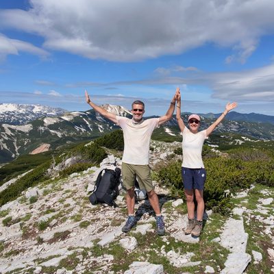 Martin und Martina stehen lachend frontal zur Kamera auf einem Karst-Gipfel und strecken triumphierend beide Arme in V-Form in den Himmel. Beide tragen helle T-Shirts, kurze Hosen, Wanderstiefel und Sonnenbrillen; ihre Wanderrucksäcke stehen neben ihnen am Boden. Sie stehen auf einer flachen Karst-Wiese mit niedrigen, immergrünen Sträuchern und vereinzelten Steinen. Im Hintergrund erstreckt sich eine weite Bergkette mit teils schneeflecken-bedeckten Felshängen und sanften, grünen Bergrücken bis zum Horizont. Strahlend blauer Himmel mit weichen weissen Wolken.