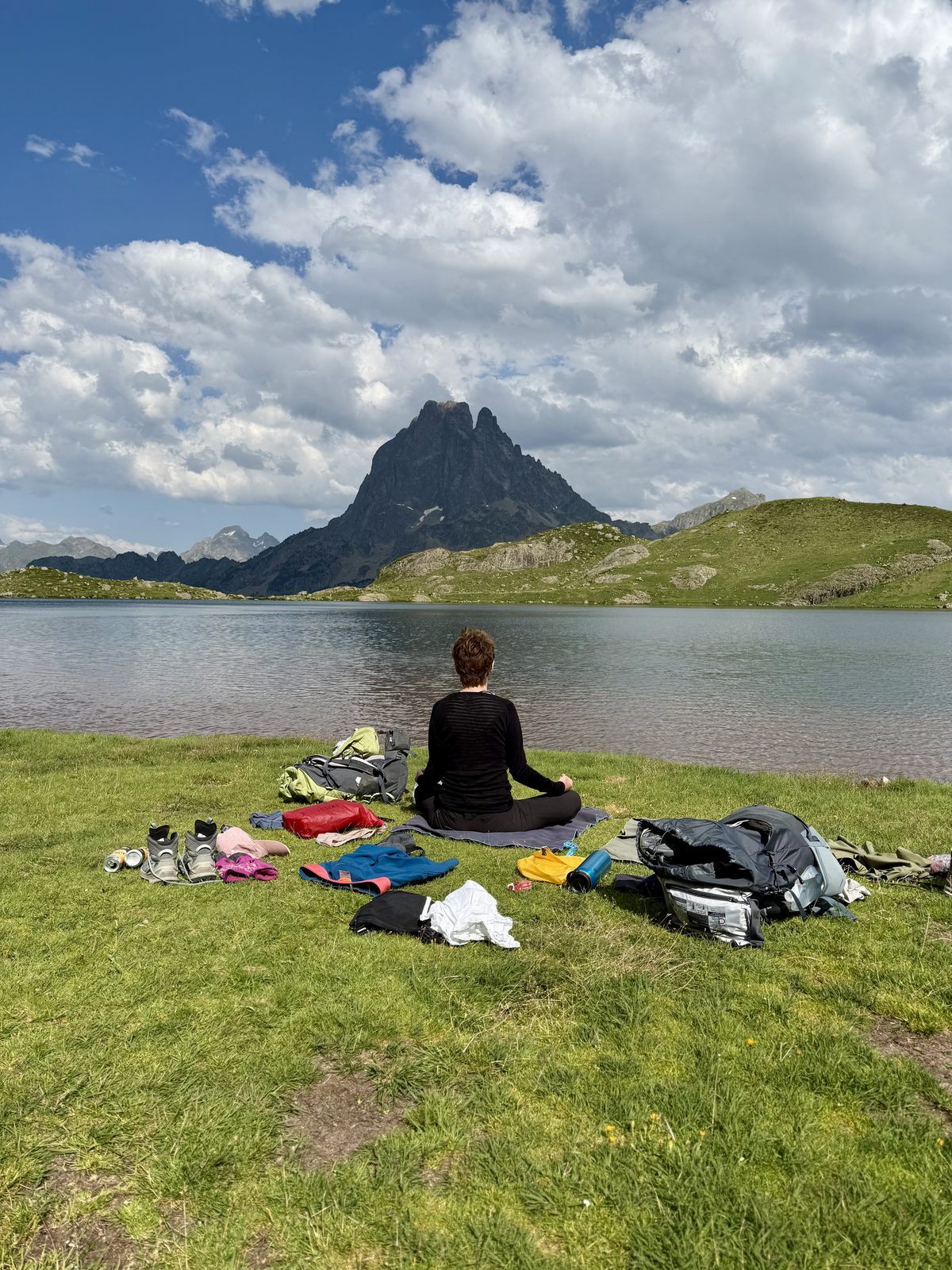 Eine Person meditiert im Schneidersitz auf einer Matte am Ufer eines ruhigen Bergsees. Ausgebreitetes Gepäck und Kleider liegen rundum auf der Wiese. Im Hintergrund ragt der markante, spitze Pic du Midi d'Ossau über dem Wasser.