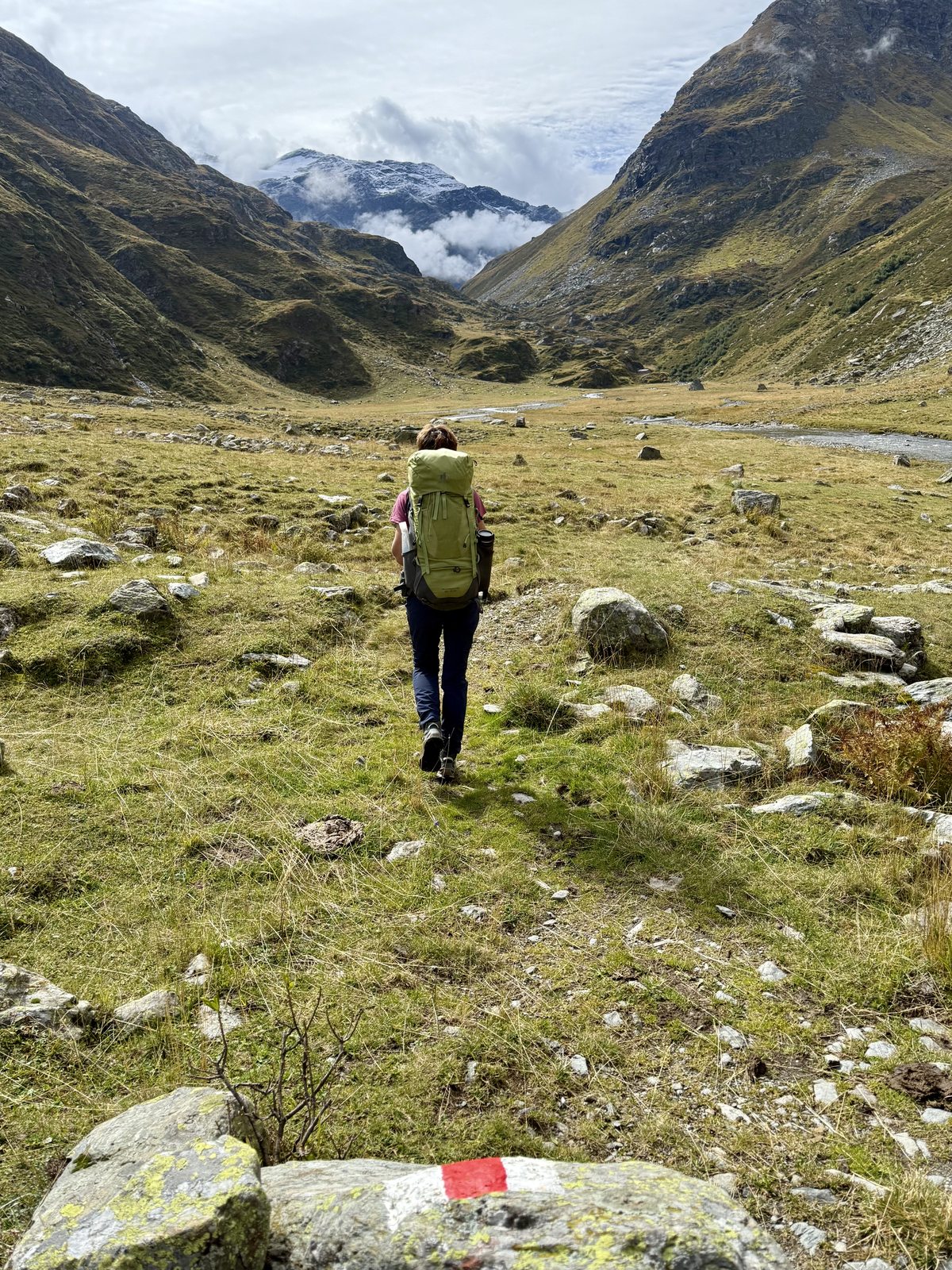 Martina geht mit grünem Rucksack allein durch ein weites, offenes Hochtal. Ein Bach fliesst rechts daneben über Steine. Im Hintergrund wolkenverhangene Felsgipfel und dunkelgraue Berge.