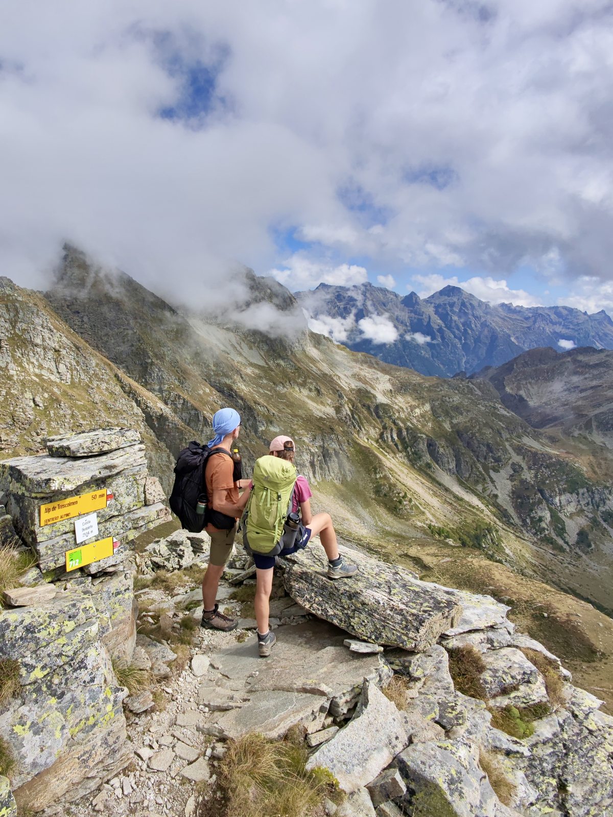 Martina und Martin stehen und sitzen an einem gelben Wegweiser auf einem Bergsattel und schauen ins Bergpanorama. Wolken ziehen um die felsigen Gipfel.