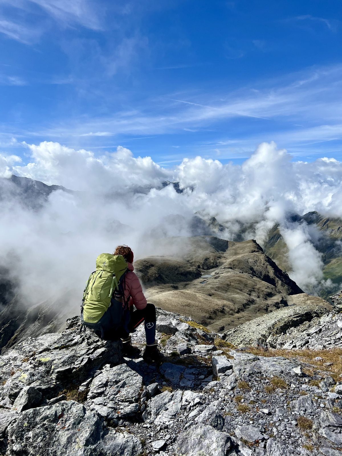Martina hockt mit grünem Rucksack und rosa Jacke auf einem Gipfelfelsen, Rücken zur Kamera, und blickt auf ein herbstliches Bergpanorama. Wolken ziehen zwischen den felsigen Gipfeln durch.