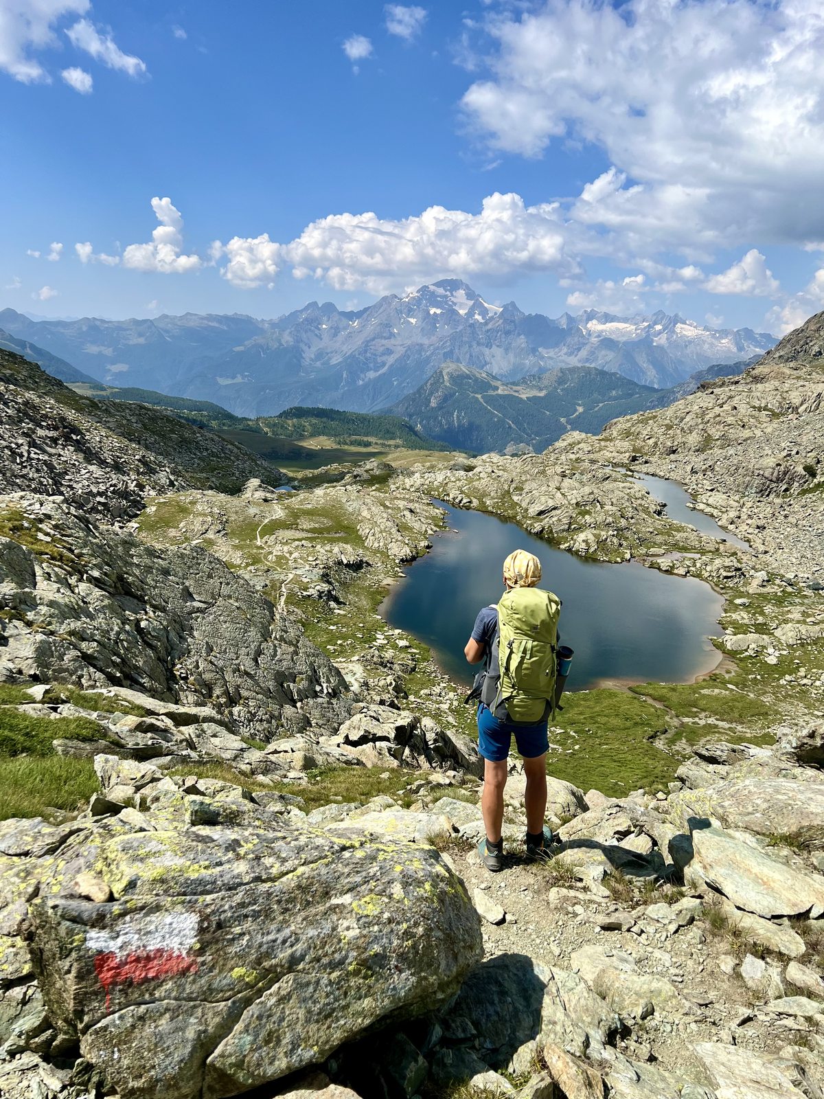 Martina steht mit grünem Rucksack auf einem Wanderpfad und schaut auf zwei kleine Bergseen in einem Hochgebirgskessel. Im Hintergrund erhebt sich ein markanter Berggipfel.