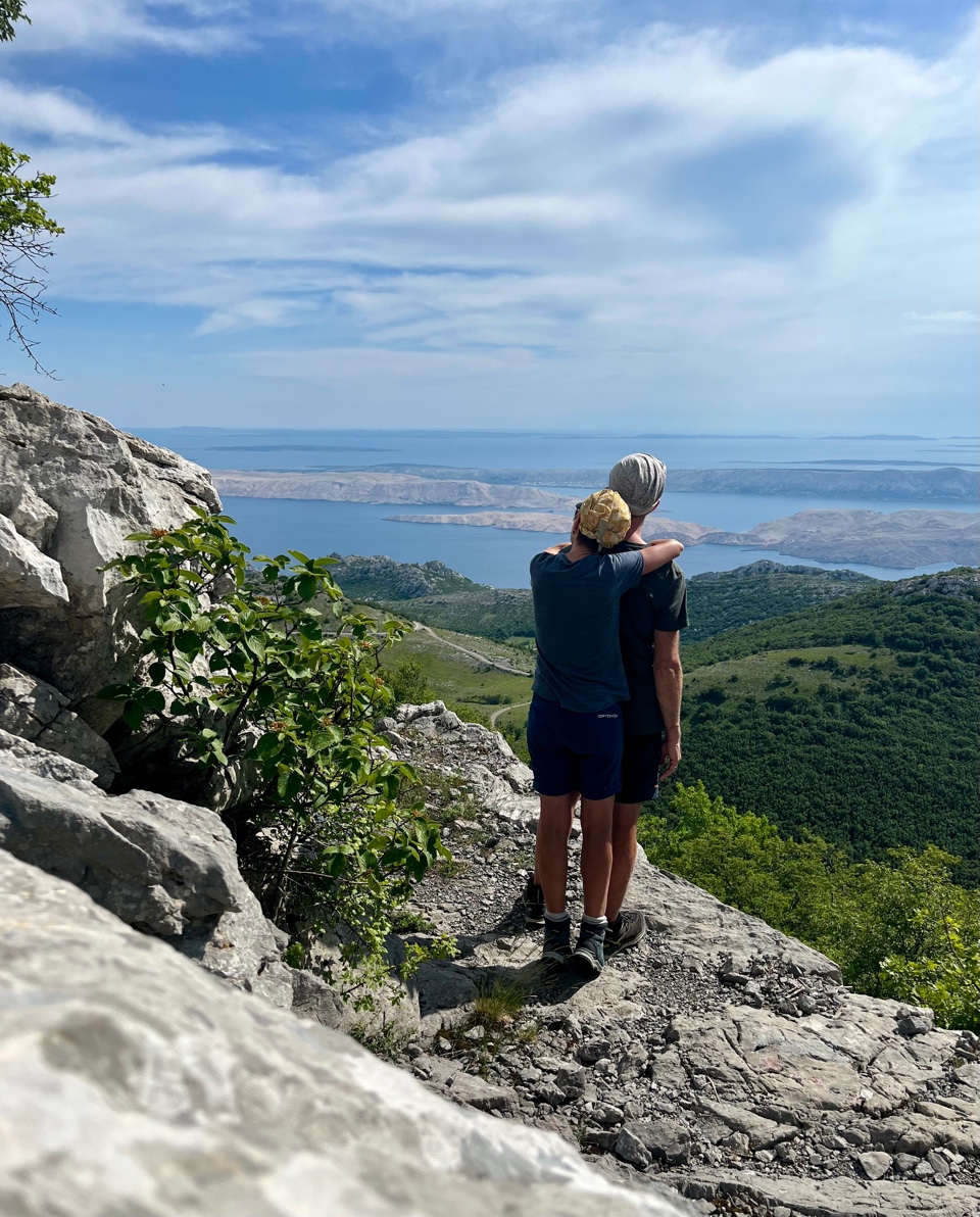 Martina und Martin stehen auf einem Felsvorsprung, er legt den Arm um ihre Schulter. Beide blicken auf die dalmatinische Küste mit Inseln im Meer darunter. Grüne Vegetation im Vordergrund.